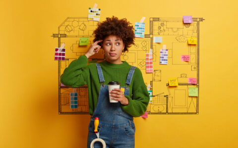 Professional hard working designer has thoughtful look, thinks about creative sketching project during coffee break, selects colors and furniture for new home interior of customers prepares house plan