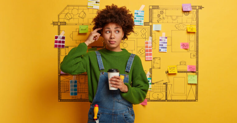 Professional hard working designer has thoughtful look, thinks about creative sketching project during coffee break, selects colors and furniture for new home interior of customers prepares house plan